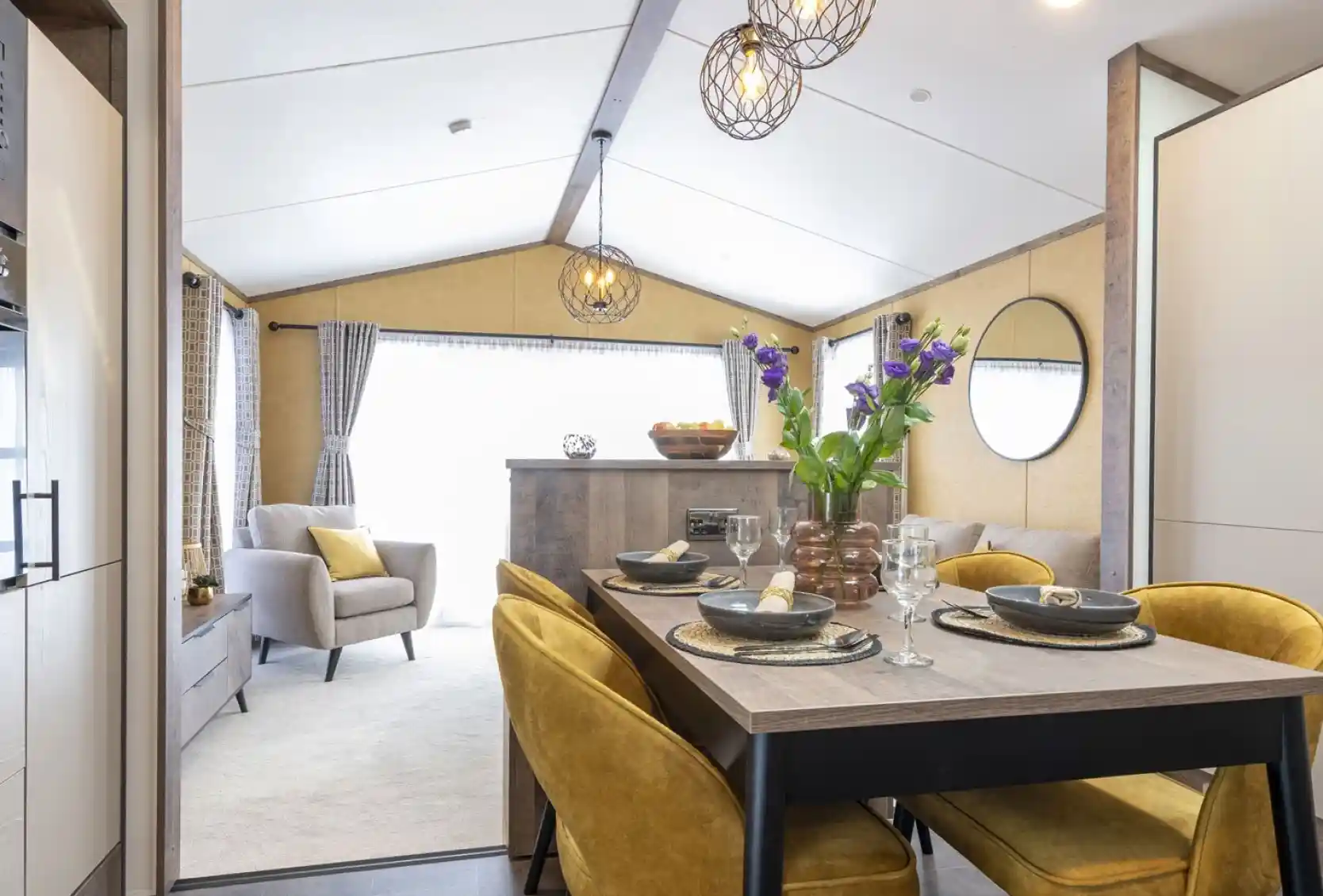 Cozy dining and living area with a wooden table, mustard-yellow chairs, and a vase of purple flowers. Light gray armchair and sofa in the background, with modern pendant lights and a round mirror on the wall. Bright and inviting space.