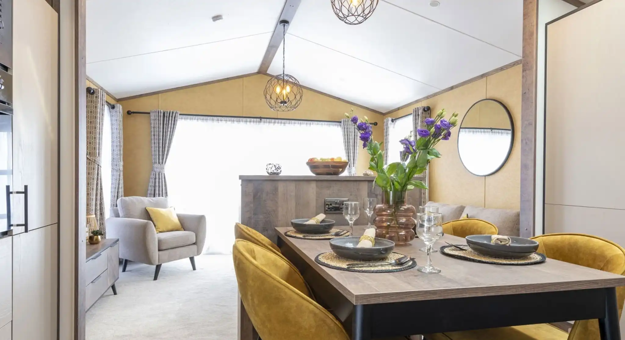 Cozy dining and living area with a wooden table, mustard-yellow chairs, and a vase of purple flowers. Light gray armchair and sofa in the background, with modern pendant lights and a round mirror on the wall. Bright and inviting space.