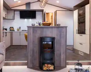 A modern kitchen and living area featuring a central wood-paneled fireplace with a cylindrical black stove. The stove has a glass front displaying a glowing fire and a compartment underneath holding logs. The kitchen has light-colored cabinets with black handles, a marble-patterned backsplash, and built-in black appliances including an oven and microwave. The floor is tiled with dark tiles, and the kitchen area is slightly elevated with two steps leading down to a carpeted living space. A decorative bowl of fruit and flowers sit on the countertop above the fireplace.