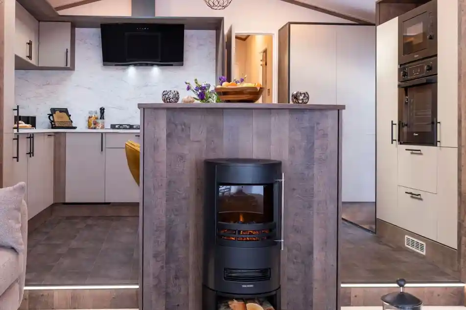 A modern kitchen and living area featuring a central wood-paneled fireplace with a cylindrical black stove. The stove has a glass front displaying a glowing fire and a compartment underneath holding logs. The kitchen has light-colored cabinets with black handles, a marble-patterned backsplash, and built-in black appliances including an oven and microwave. The floor is tiled with dark tiles, and the kitchen area is slightly elevated with two steps leading down to a carpeted living space. A decorative bowl of fruit and flowers sit on the countertop above the fireplace.