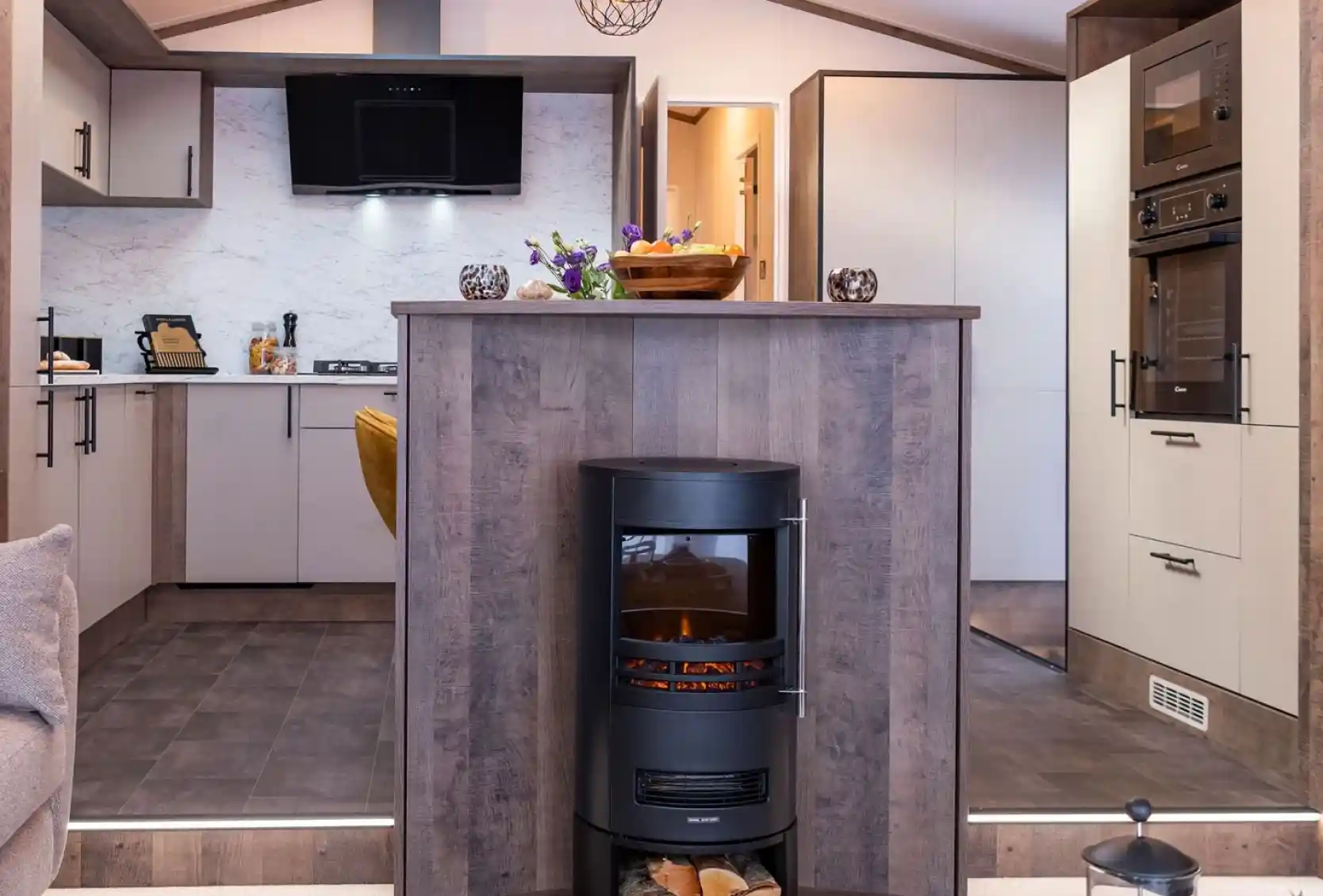 A modern kitchen and living area featuring a central wood-paneled fireplace with a cylindrical black stove. The stove has a glass front displaying a glowing fire and a compartment underneath holding logs. The kitchen has light-colored cabinets with black handles, a marble-patterned backsplash, and built-in black appliances including an oven and microwave. The floor is tiled with dark tiles, and the kitchen area is slightly elevated with two steps leading down to a carpeted living space. A decorative bowl of fruit and flowers sit on the countertop above the fireplace.