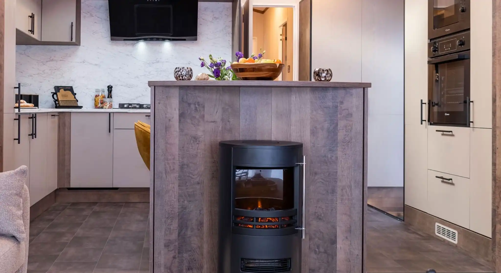 A modern kitchen and living area featuring a central wood-paneled fireplace with a cylindrical black stove. The stove has a glass front displaying a glowing fire and a compartment underneath holding logs. The kitchen has light-colored cabinets with black handles, a marble-patterned backsplash, and built-in black appliances including an oven and microwave. The floor is tiled with dark tiles, and the kitchen area is slightly elevated with two steps leading down to a carpeted living space. A decorative bowl of fruit and flowers sit on the countertop above the fireplace.