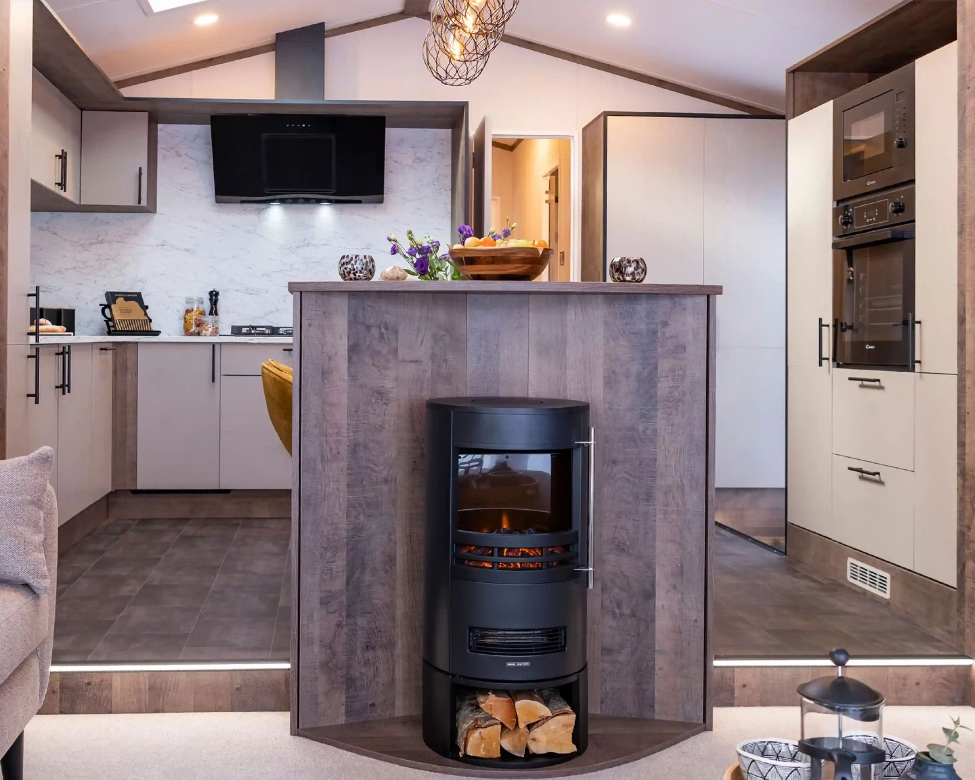 A modern kitchen and living area featuring a central wood-paneled fireplace with a cylindrical black stove. The stove has a glass front displaying a glowing fire and a compartment underneath holding logs. The kitchen has light-colored cabinets with black handles, a marble-patterned backsplash, and built-in black appliances including an oven and microwave. The floor is tiled with dark tiles, and the kitchen area is slightly elevated with two steps leading down to a carpeted living space. A decorative bowl of fruit and flowers sit on the countertop above the fireplace.