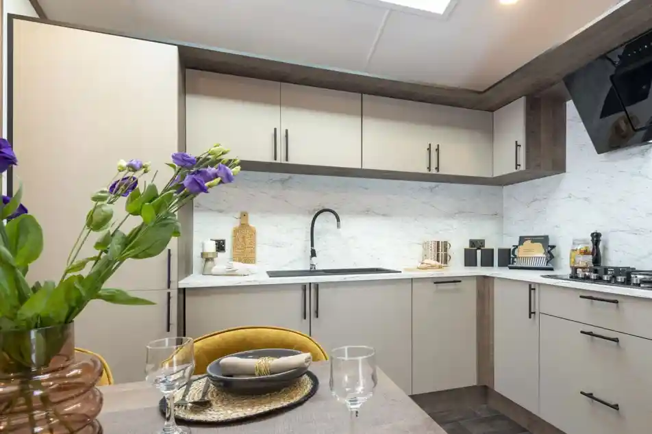 Modern kitchen with beige cabinets and black handles, featuring a marble-look backsplash and countertop. A black faucet is centered above the sink. A gas stove is visible on the right. In the foreground, a dining table is set with dark plates, beige napkins with gold rings, clear wine glasses, and a brown vase holding purple flowers. The kitchen is well-lit with natural light from a skylight above.