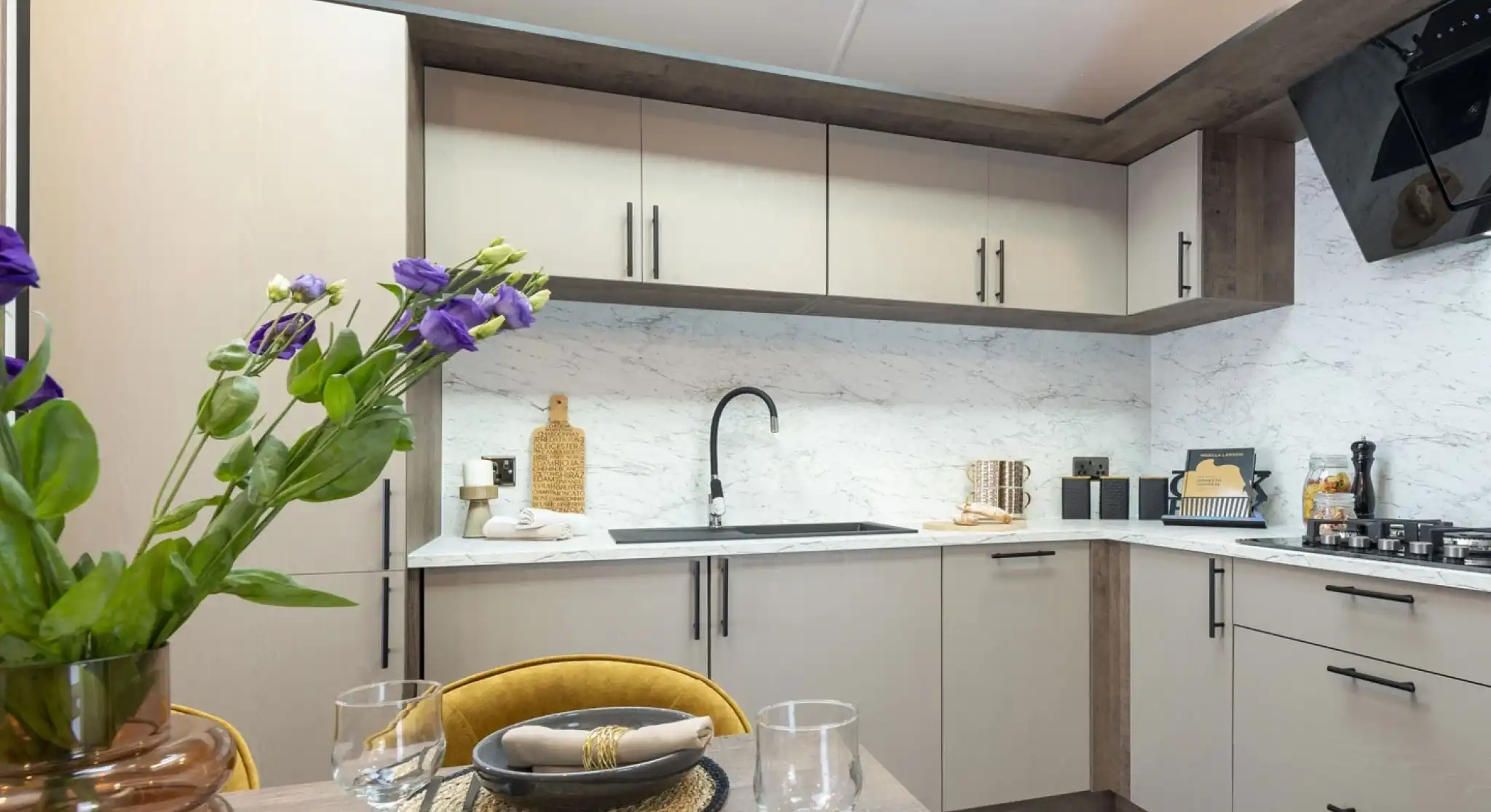 Modern kitchen with beige cabinets and black handles, featuring a marble-look backsplash and countertop. A black faucet is centered above the sink. A gas stove is visible on the right. In the foreground, a dining table is set with dark plates, beige napkins with gold rings, clear wine glasses, and a brown vase holding purple flowers. The kitchen is well-lit with natural light from a skylight above.