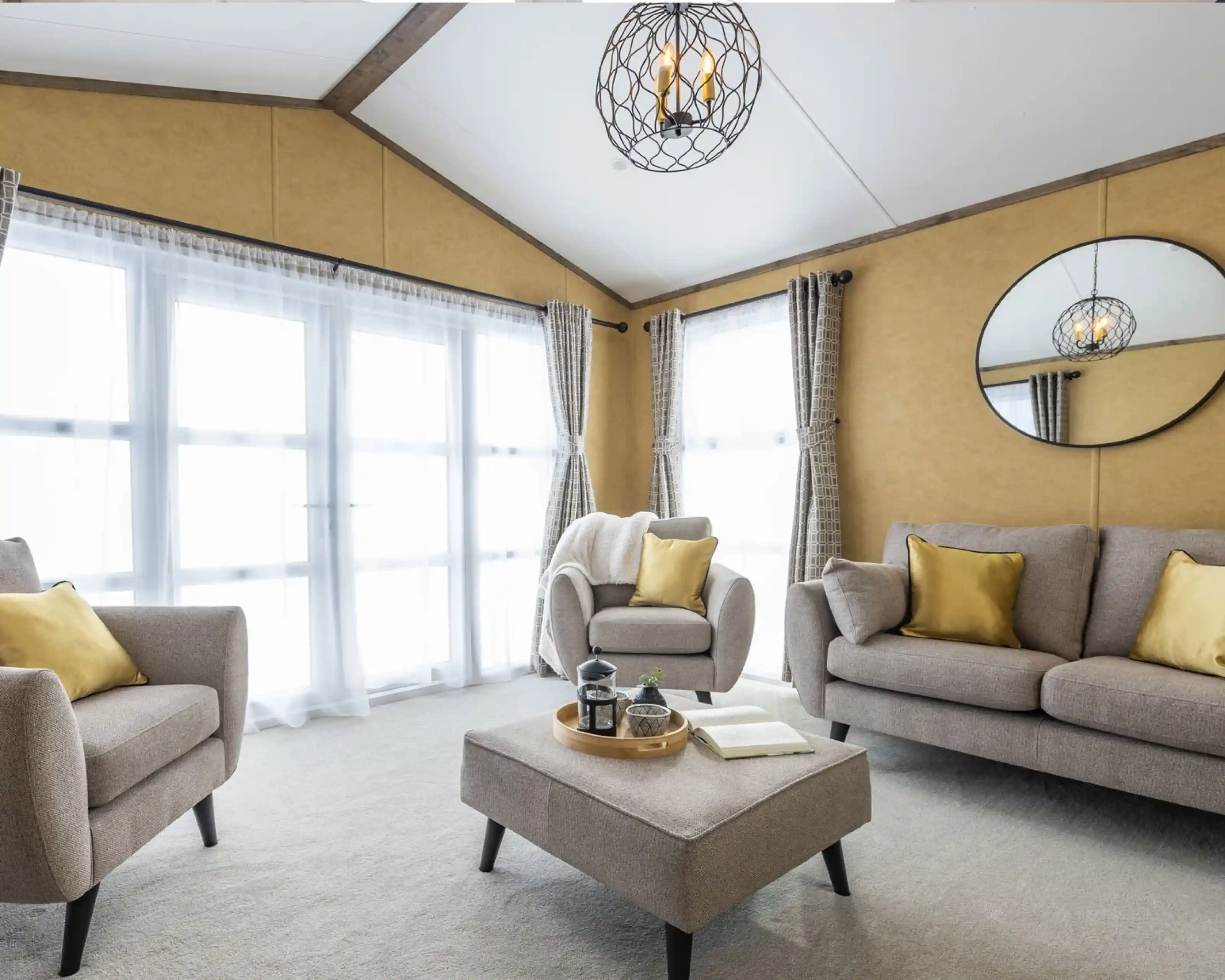 This is a bright and cozy living room featuring neutral tones with pops of yellow. It has large windows covered with sheer white curtains, allowing plenty of natural light to flood the space. The furniture includes a beige sofa, two armchairs, and a matching ottoman in the center. Yellow throw pillows add a vibrant touch to the seating. A circular mirror hangs on the mustard-colored wall, reflecting the room's light fixture, which is a decorative pendant with exposed bulbs enclosed in a wireframe. The overall style is modern and inviting, with a warm, earthy color palette.
