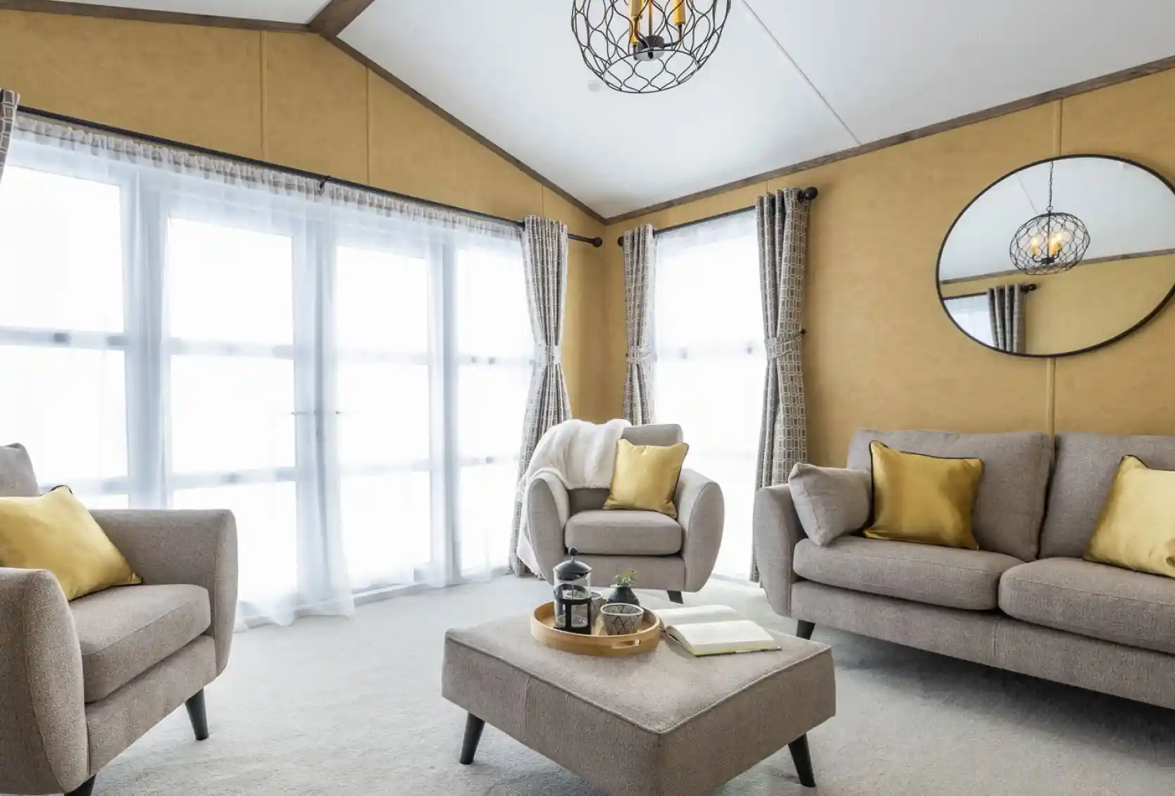 This is a bright and cozy living room featuring neutral tones with pops of yellow. It has large windows covered with sheer white curtains, allowing plenty of natural light to flood the space. The furniture includes a beige sofa, two armchairs, and a matching ottoman in the center. Yellow throw pillows add a vibrant touch to the seating. A circular mirror hangs on the mustard-colored wall, reflecting the room's light fixture, which is a decorative pendant with exposed bulbs enclosed in a wireframe. The overall style is modern and inviting, with a warm, earthy color palette.