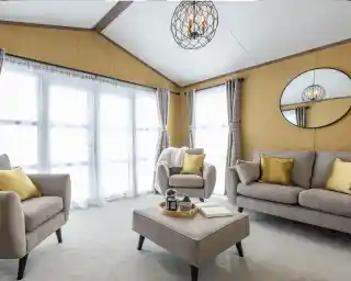 This is a bright and cozy living room featuring neutral tones with pops of yellow. It has large windows covered with sheer white curtains, allowing plenty of natural light to flood the space. The furniture includes a beige sofa, two armchairs, and a matching ottoman in the center. Yellow throw pillows add a vibrant touch to the seating. A circular mirror hangs on the mustard-colored wall, reflecting the room's light fixture, which is a decorative pendant with exposed bulbs enclosed in a wireframe. The overall style is modern and inviting, with a warm, earthy color palette.