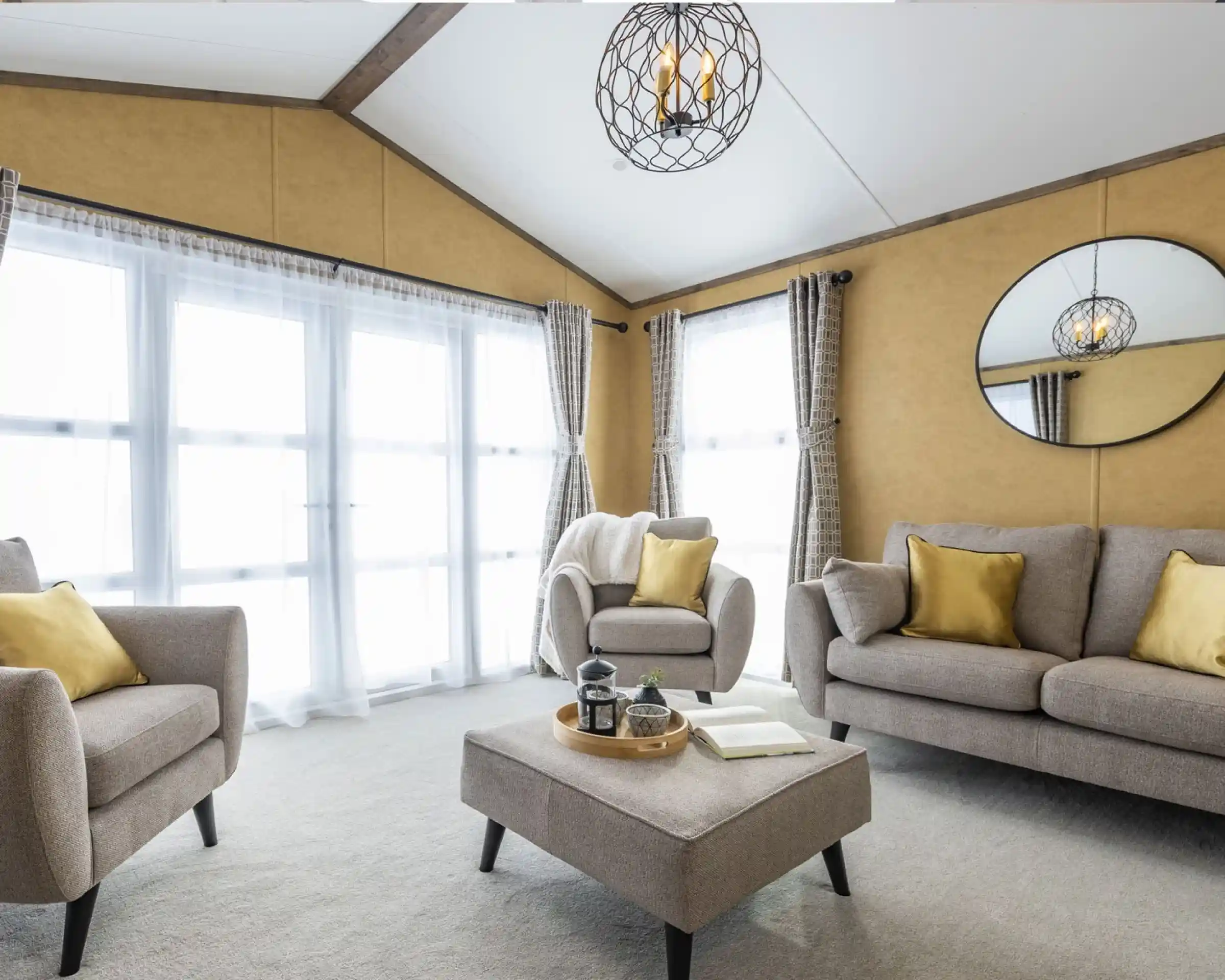 This is a bright and cozy living room featuring neutral tones with pops of yellow. It has large windows covered with sheer white curtains, allowing plenty of natural light to flood the space. The furniture includes a beige sofa, two armchairs, and a matching ottoman in the center. Yellow throw pillows add a vibrant touch to the seating. A circular mirror hangs on the mustard-colored wall, reflecting the room's light fixture, which is a decorative pendant with exposed bulbs enclosed in a wireframe. The overall style is modern and inviting, with a warm, earthy color palette.