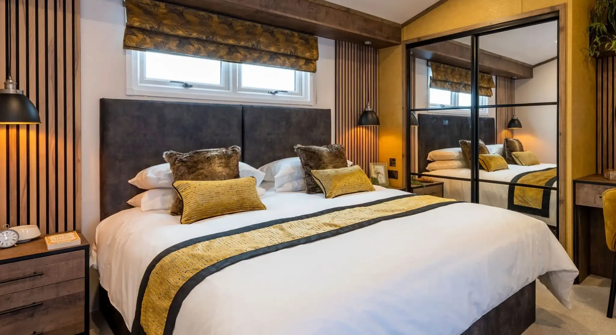 A modern, cozy bedroom with a large double bed featuring white bedding and decorative yellow and brown pillows and a matching throw. The bed has a dark upholstered headboard against a white wall with vertical wooden slats on either side. Above the headboard, there are two small rectangular windows with a yellow patterned Roman blind. On both sides of the bed, black wall-mounted reading lamps provide warm lighting. To the right, there is a large mirrored wardrobe with sliding doors reflecting the bed and part of the room. The room has warm yellow walls and a beige carpet, creating a welcoming and comfortable atmosphere.
