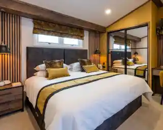 A modern, cozy bedroom with a large double bed featuring white bedding and decorative yellow and brown pillows and a matching throw. The bed has a dark upholstered headboard against a white wall with vertical wooden slats on either side. Above the headboard, there are two small rectangular windows with a yellow patterned Roman blind. On both sides of the bed, black wall-mounted reading lamps provide warm lighting. To the right, there is a large mirrored wardrobe with sliding doors reflecting the bed and part of the room. The room has warm yellow walls and a beige carpet, creating a welcoming and comfortable atmosphere.