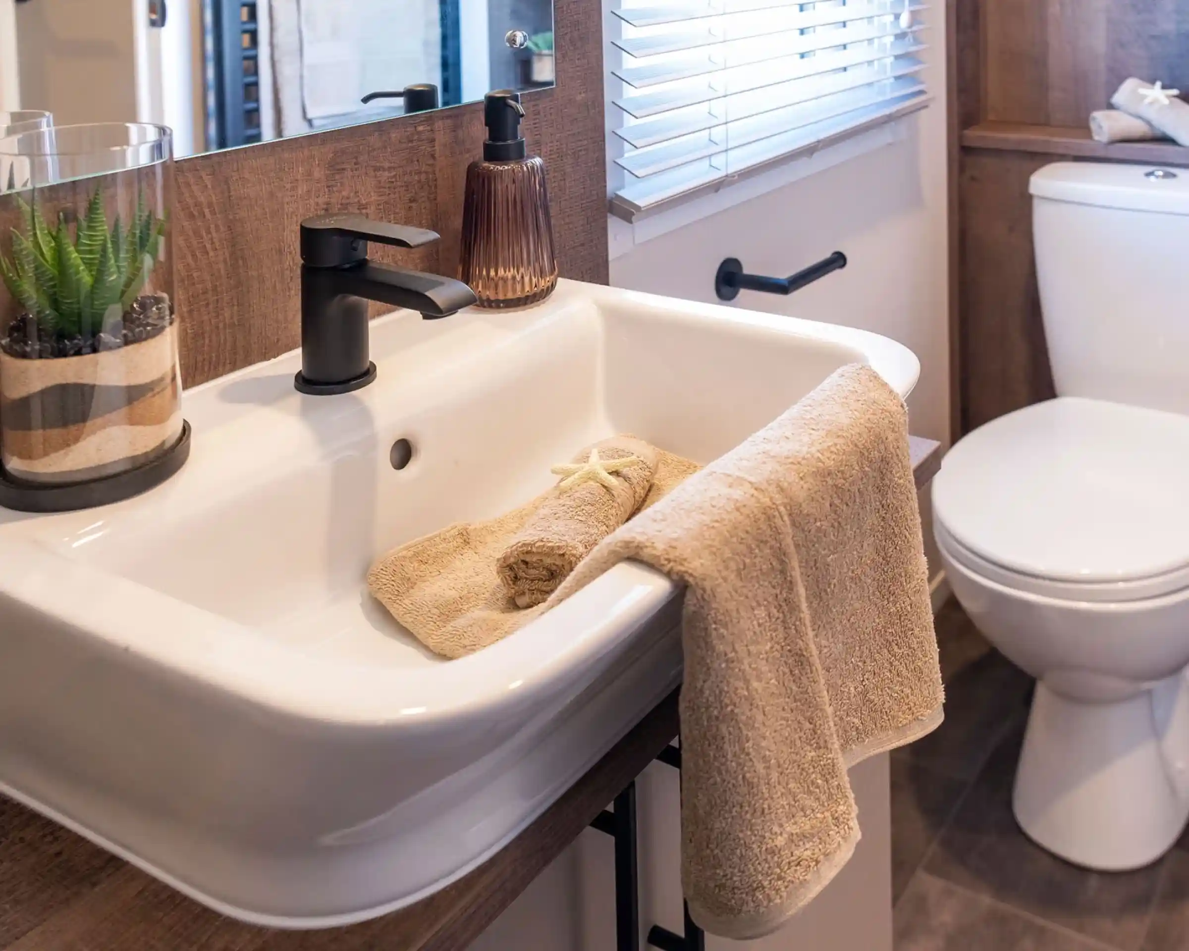 This image shows a modern, clean bathroom sink area. The sink is white with a matte black faucet. There is a beige hand towel hanging on the sink edge, and another rolled-up beige towel inside the sink, decorated with a small starfish. Next to the faucet is a brownish glass soap dispenser. On the left side of the sink, there's a glass container holding a green succulent plant with layered sand. In the background, there is a window with white blinds and a white toilet with a towel and a starfish on the tank lid. The overall design has a natural, beach-inspired theme with neutral tones and wood accents