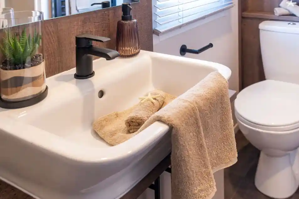 This image shows a modern, clean bathroom sink area. The sink is white with a matte black faucet. There is a beige hand towel hanging on the sink edge, and another rolled-up beige towel inside the sink, decorated with a small starfish. Next to the faucet is a brownish glass soap dispenser. On the left side of the sink, there's a glass container holding a green succulent plant with layered sand. In the background, there is a window with white blinds and a white toilet with a towel and a starfish on the tank lid. The overall design has a natural, beach-inspired theme with neutral tones and wood accents