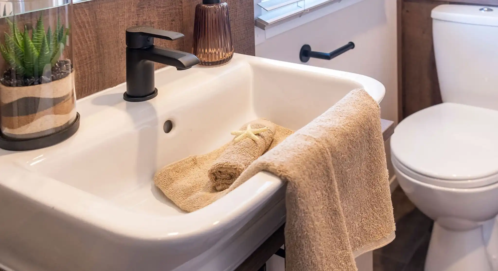 This image shows a modern, clean bathroom sink area. The sink is white with a matte black faucet. There is a beige hand towel hanging on the sink edge, and another rolled-up beige towel inside the sink, decorated with a small starfish. Next to the faucet is a brownish glass soap dispenser. On the left side of the sink, there's a glass container holding a green succulent plant with layered sand. In the background, there is a window with white blinds and a white toilet with a towel and a starfish on the tank lid. The overall design has a natural, beach-inspired theme with neutral tones and wood accents