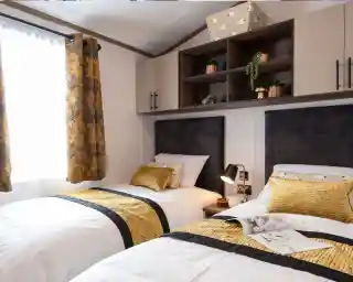This image shows a cozy twin bedroom with two single beds. Each bed has white bedding with a mustard-yellow and black patterned runner and matching yellow pillows. There are dark upholstered headboards behind each bed. Between the beds is a small bedside table with a modern black and gold lamp, a cup, and a small decorative item. Above the beds, there are wall-mounted cabinets with both closed storage and open shelving, decorated with small potted plants and a golden mushroom ornament. The room has a window with mustard-yellow curtains featuring a leafy pattern, letting in natural light.