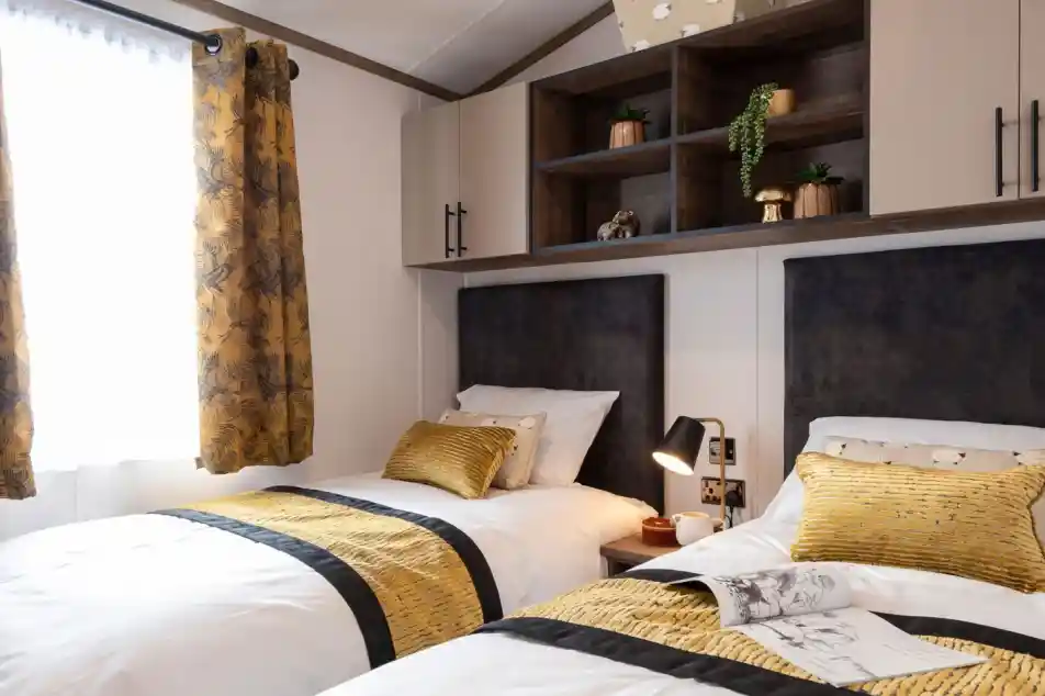 This image shows a cozy twin bedroom with two single beds. Each bed has white bedding with a mustard-yellow and black patterned runner and matching yellow pillows. There are dark upholstered headboards behind each bed. Between the beds is a small bedside table with a modern black and gold lamp, a cup, and a small decorative item. Above the beds, there are wall-mounted cabinets with both closed storage and open shelving, decorated with small potted plants and a golden mushroom ornament. The room has a window with mustard-yellow curtains featuring a leafy pattern, letting in natural light.