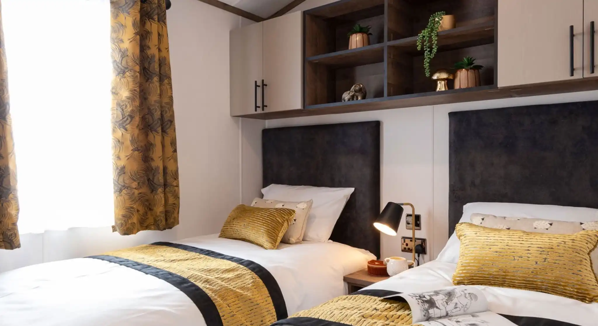 This image shows a cozy twin bedroom with two single beds. Each bed has white bedding with a mustard-yellow and black patterned runner and matching yellow pillows. There are dark upholstered headboards behind each bed. Between the beds is a small bedside table with a modern black and gold lamp, a cup, and a small decorative item. Above the beds, there are wall-mounted cabinets with both closed storage and open shelving, decorated with small potted plants and a golden mushroom ornament. The room has a window with mustard-yellow curtains featuring a leafy pattern, letting in natural light.