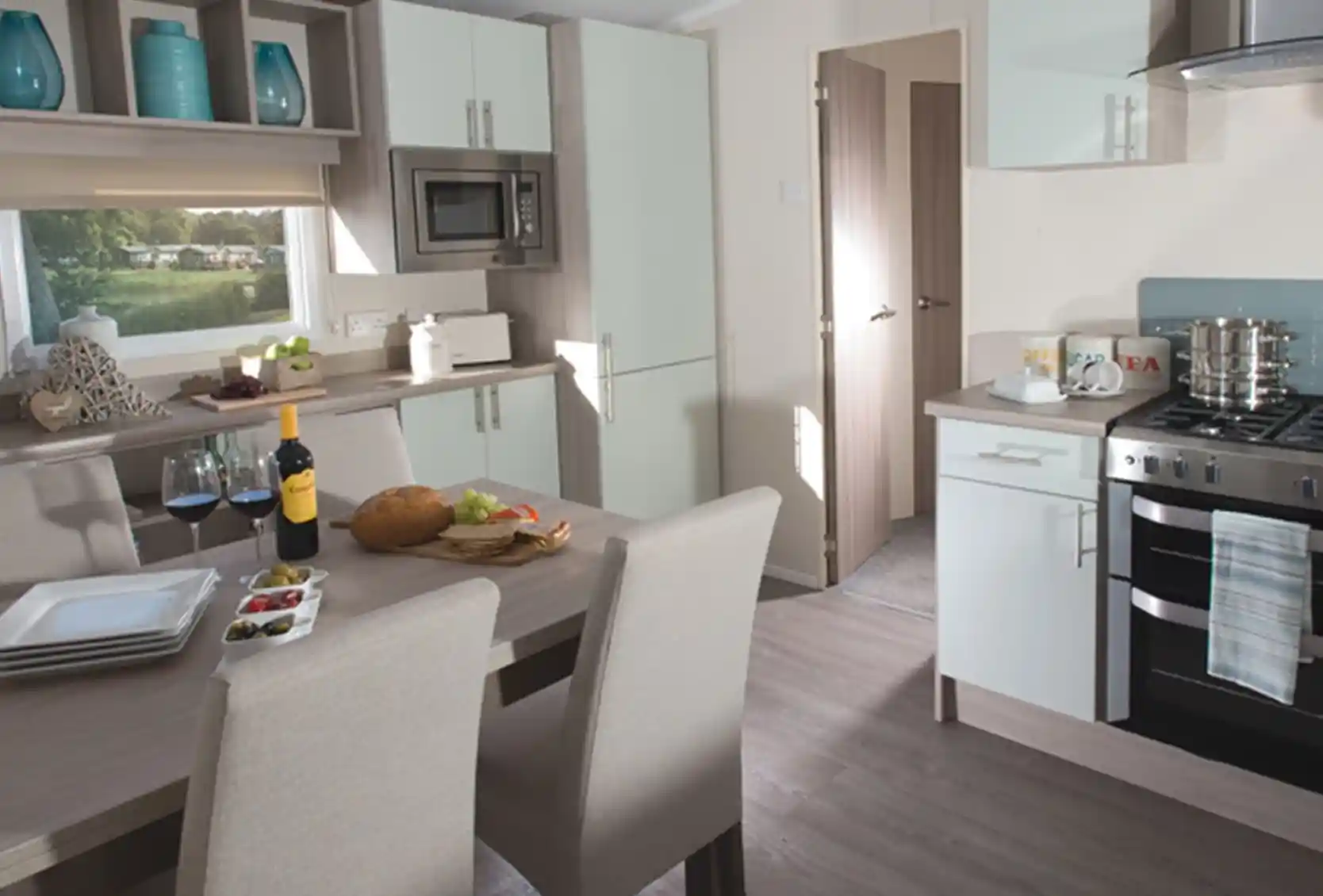 Modern holiday home kitchen and dining area featuring a wooden dining table set for four, a built-in oven with hob, light-toned cabinetry, integrated microwave, and large window with a view of a green outdoor area.