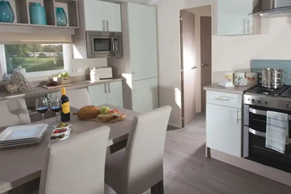 Modern holiday home kitchen and dining area featuring a wooden dining table set for four, a built-in oven with hob, light-toned cabinetry, integrated microwave, and large window with a view of a green outdoor area.