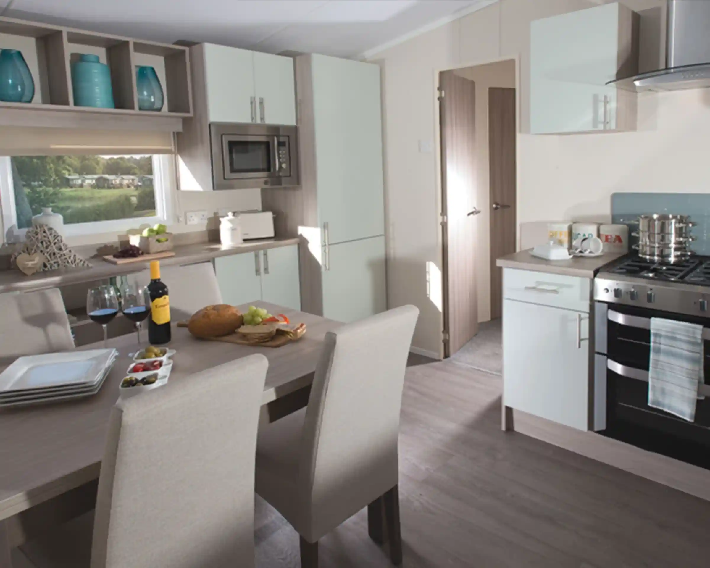 Modern holiday home kitchen and dining area featuring a wooden dining table set for four, a built-in oven with hob, light-toned cabinetry, integrated microwave, and large window with a view of a green outdoor area.
