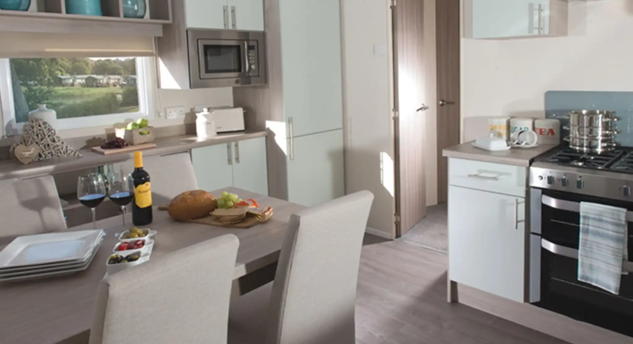 Modern holiday home kitchen and dining area featuring a wooden dining table set for four, a built-in oven with hob, light-toned cabinetry, integrated microwave, and large window with a view of a green outdoor area.