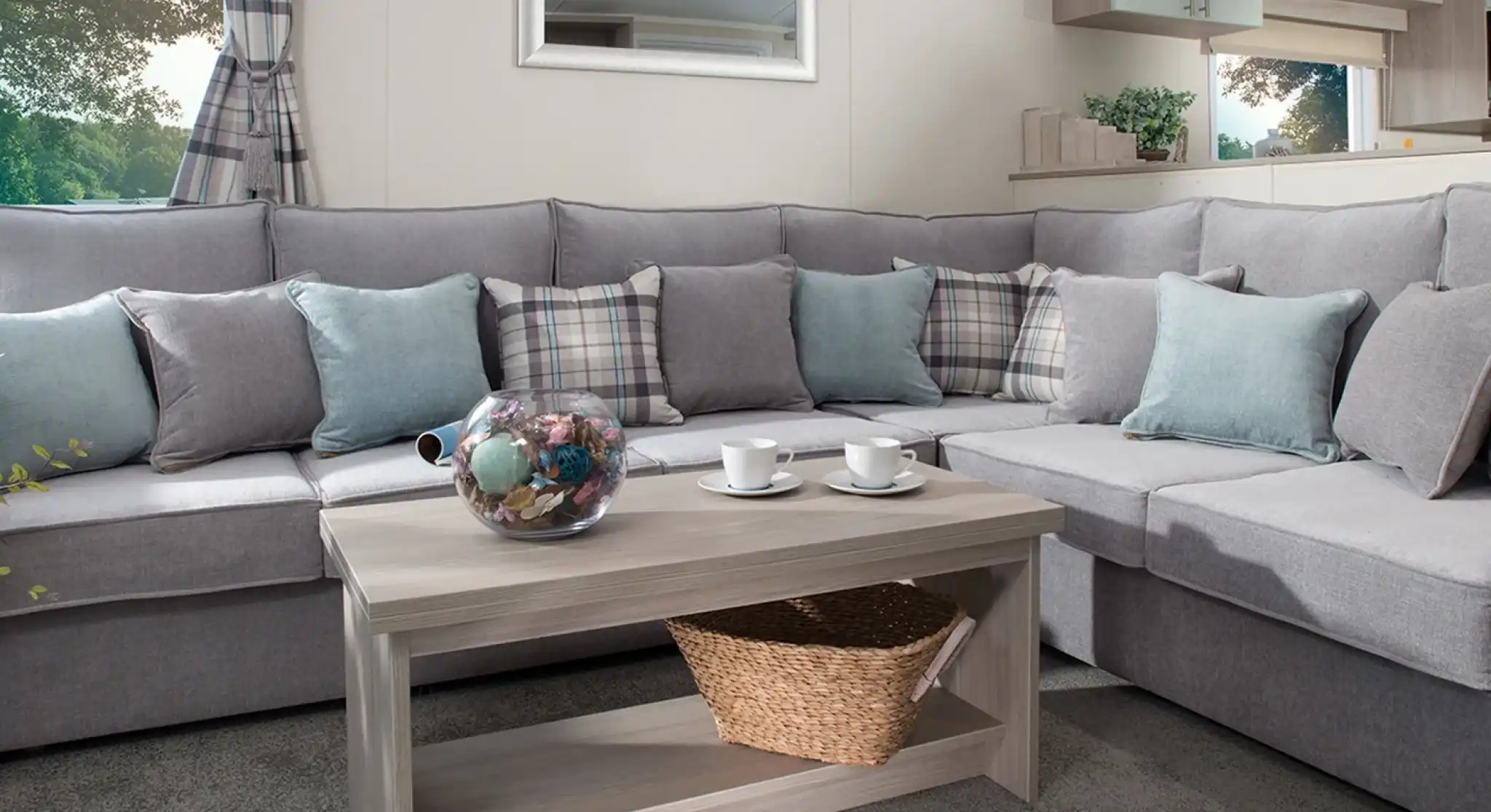Modern holiday home lounge with a large grey L-shaped sofa, pastel blue and plaid cushions, a wooden coffee table with a wicker basket, and a window with checked curtains overlooking greenery.