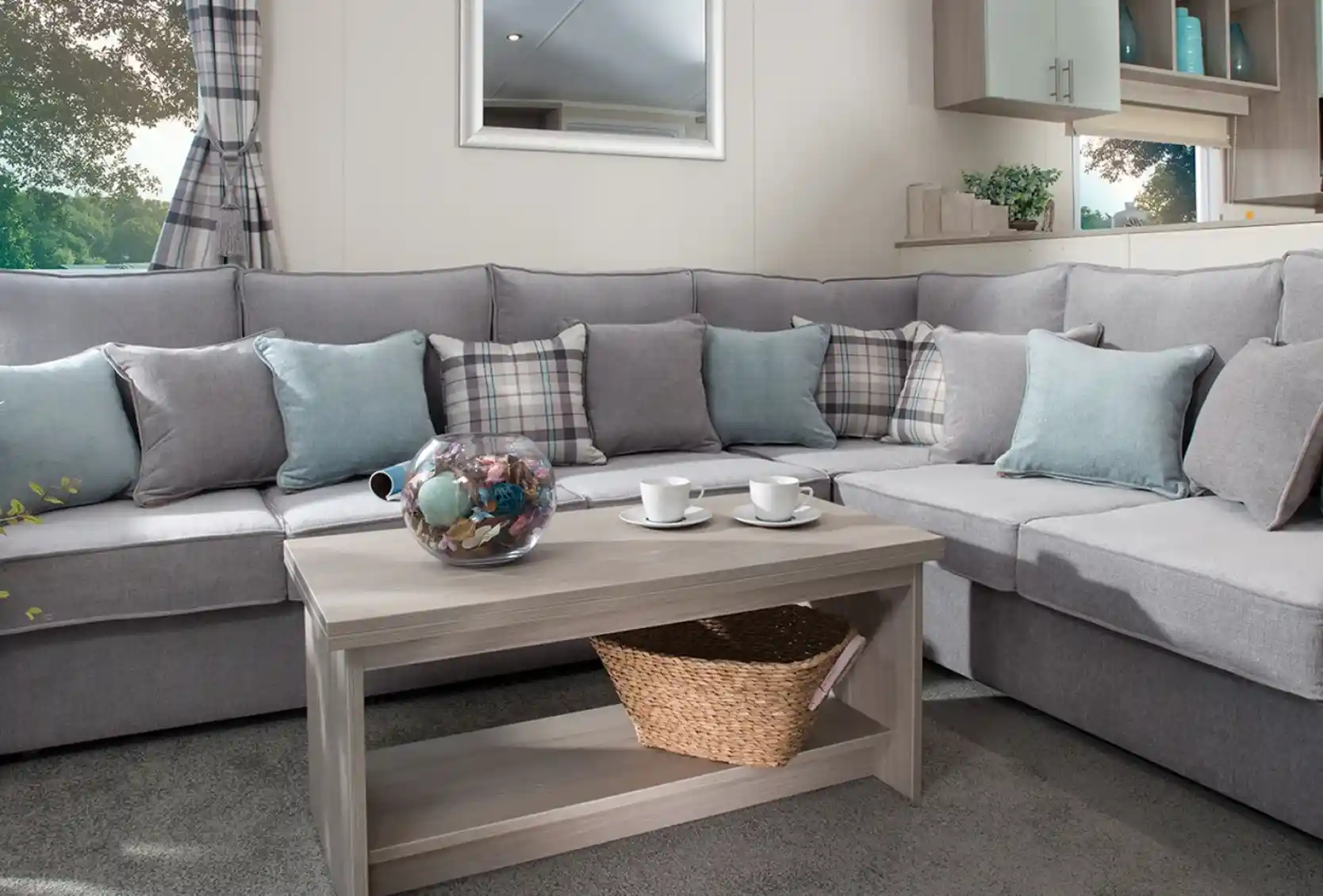 Modern holiday home lounge with a large grey L-shaped sofa, pastel blue and plaid cushions, a wooden coffee table with a wicker basket, and a window with checked curtains overlooking greenery.