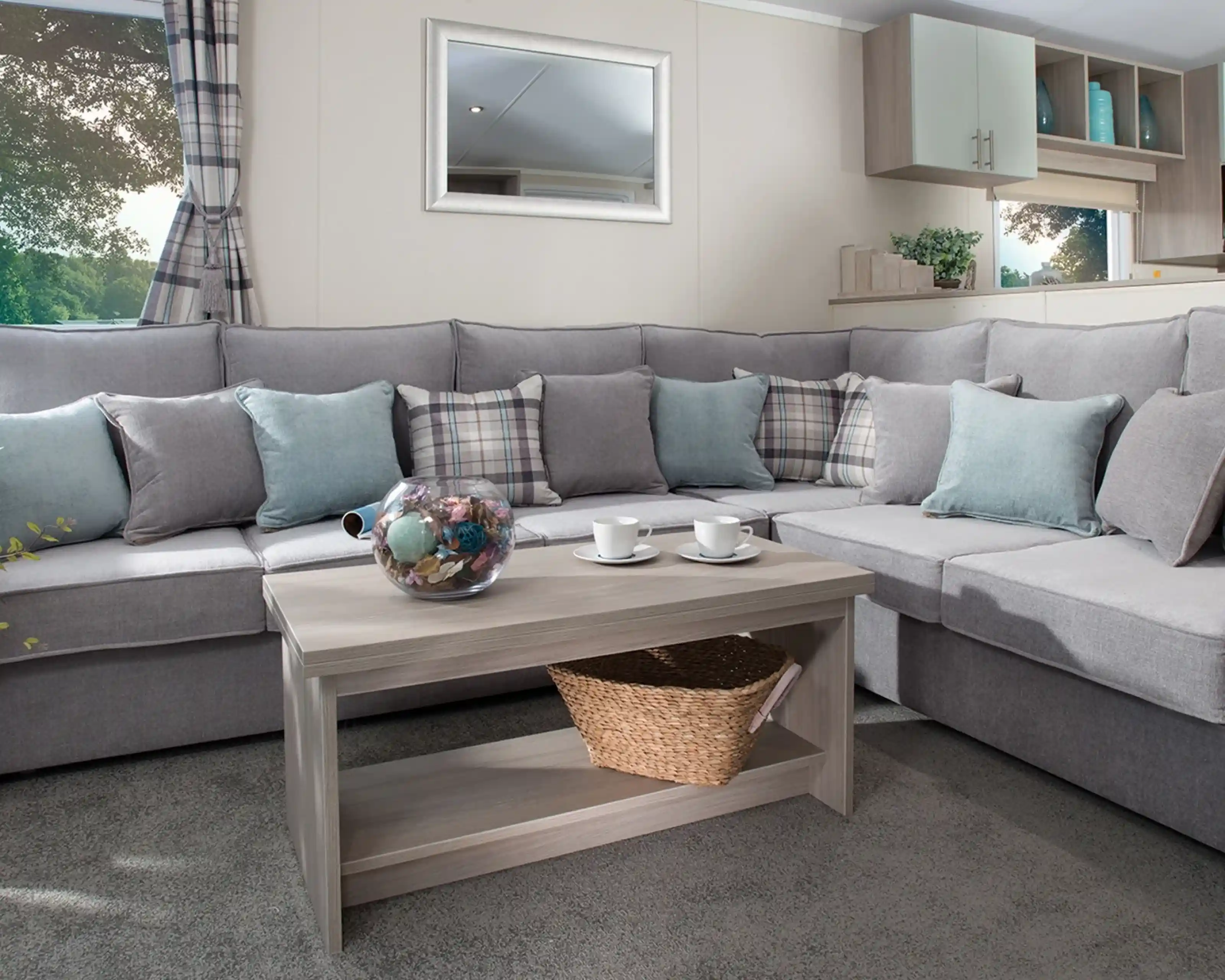 Modern holiday home lounge with a large grey L-shaped sofa, pastel blue and plaid cushions, a wooden coffee table with a wicker basket, and a window with checked curtains overlooking greenery.