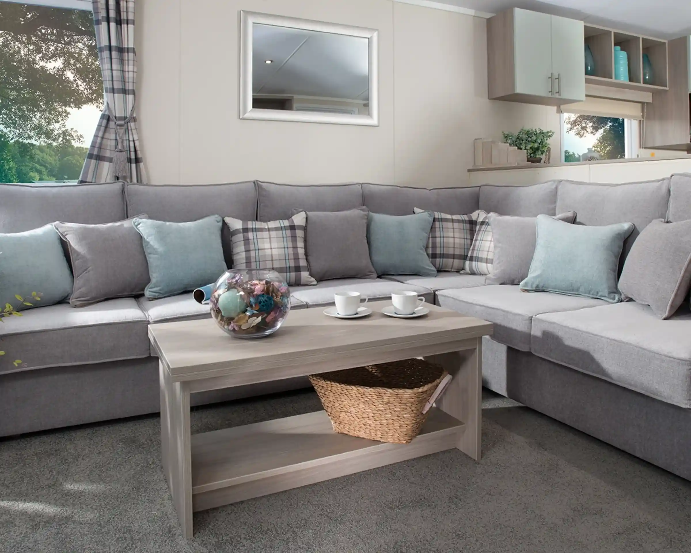 Modern holiday home lounge with a large grey L-shaped sofa, pastel blue and plaid cushions, a wooden coffee table with a wicker basket, and a window with checked curtains overlooking greenery.