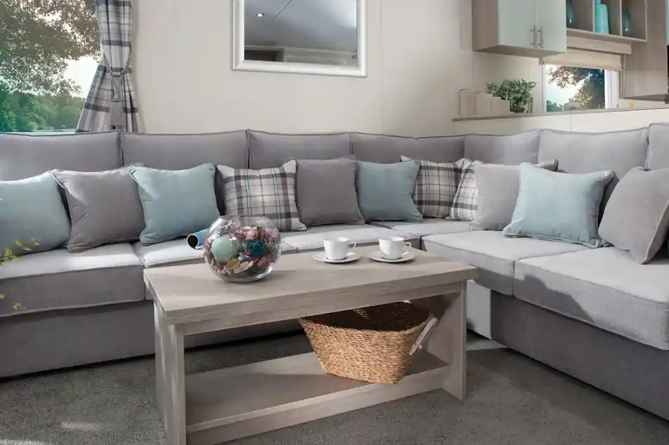 Modern holiday home lounge with a large grey L-shaped sofa, pastel blue and plaid cushions, a wooden coffee table with a wicker basket, and a window with checked curtains overlooking greenery.