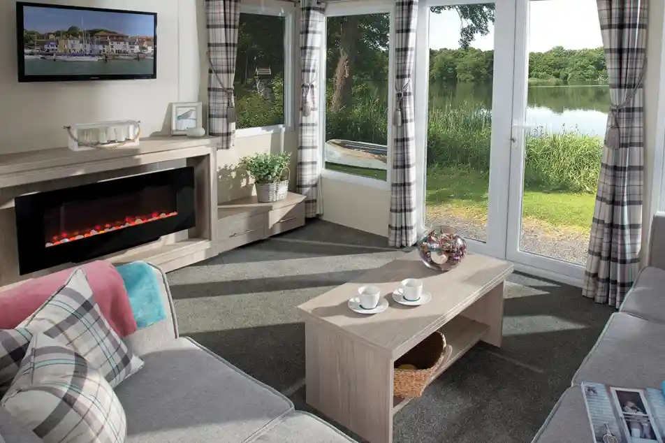 Bright holiday home lounge featuring a large corner sofa, electric fireplace, wall-mounted TV, coffee table with cups, and patio doors opening to a scenic lake view. Room is decorated with plaid curtains and soft furnishings in neutral tones.