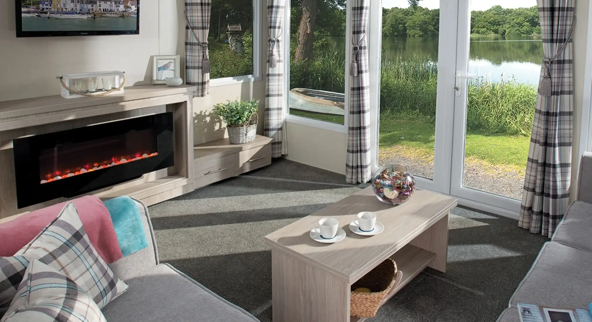 Bright holiday home lounge featuring a large corner sofa, electric fireplace, wall-mounted TV, coffee table with cups, and patio doors opening to a scenic lake view. Room is decorated with plaid curtains and soft furnishings in neutral tones.