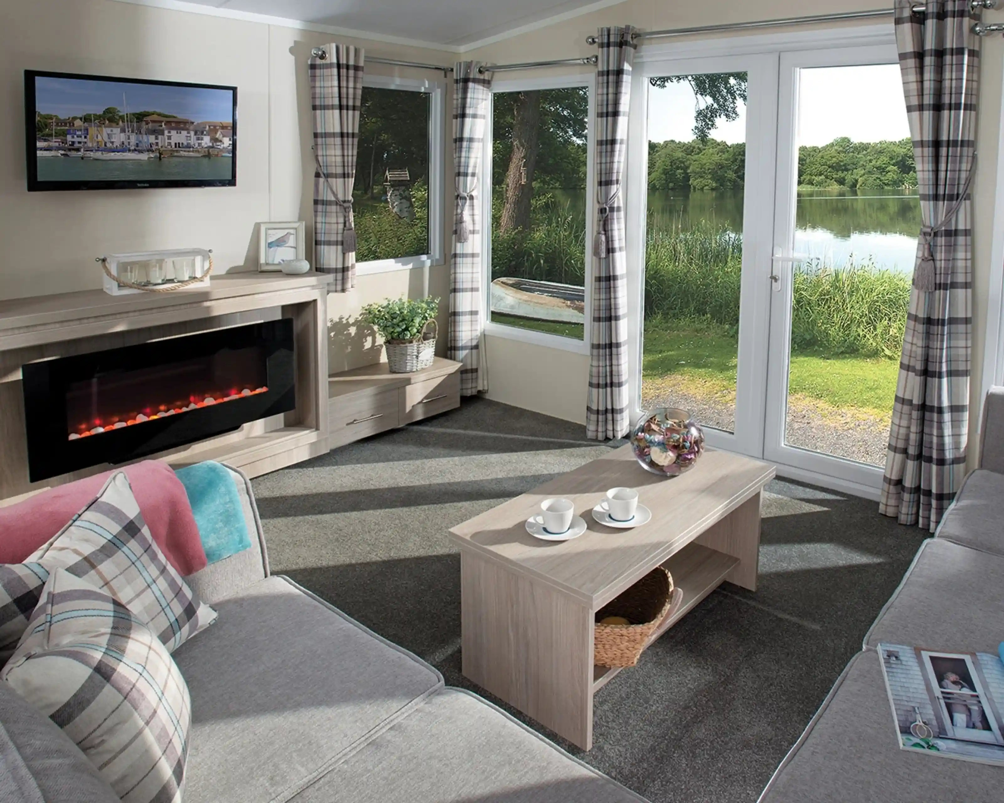 Bright holiday home lounge featuring a large corner sofa, electric fireplace, wall-mounted TV, coffee table with cups, and patio doors opening to a scenic lake view. Room is decorated with plaid curtains and soft furnishings in neutral tones.