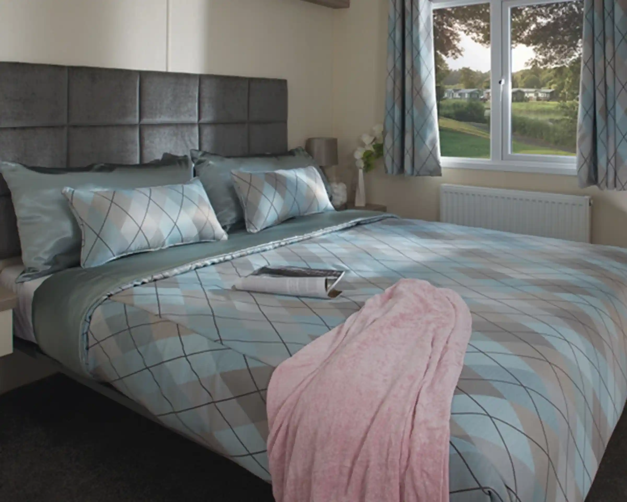 Cosy bedroom with a large bed featuring blue and gray argyle-patterned bedding, a pink blanket, and a magazine. Soft light streams through a window overlooking greenery.