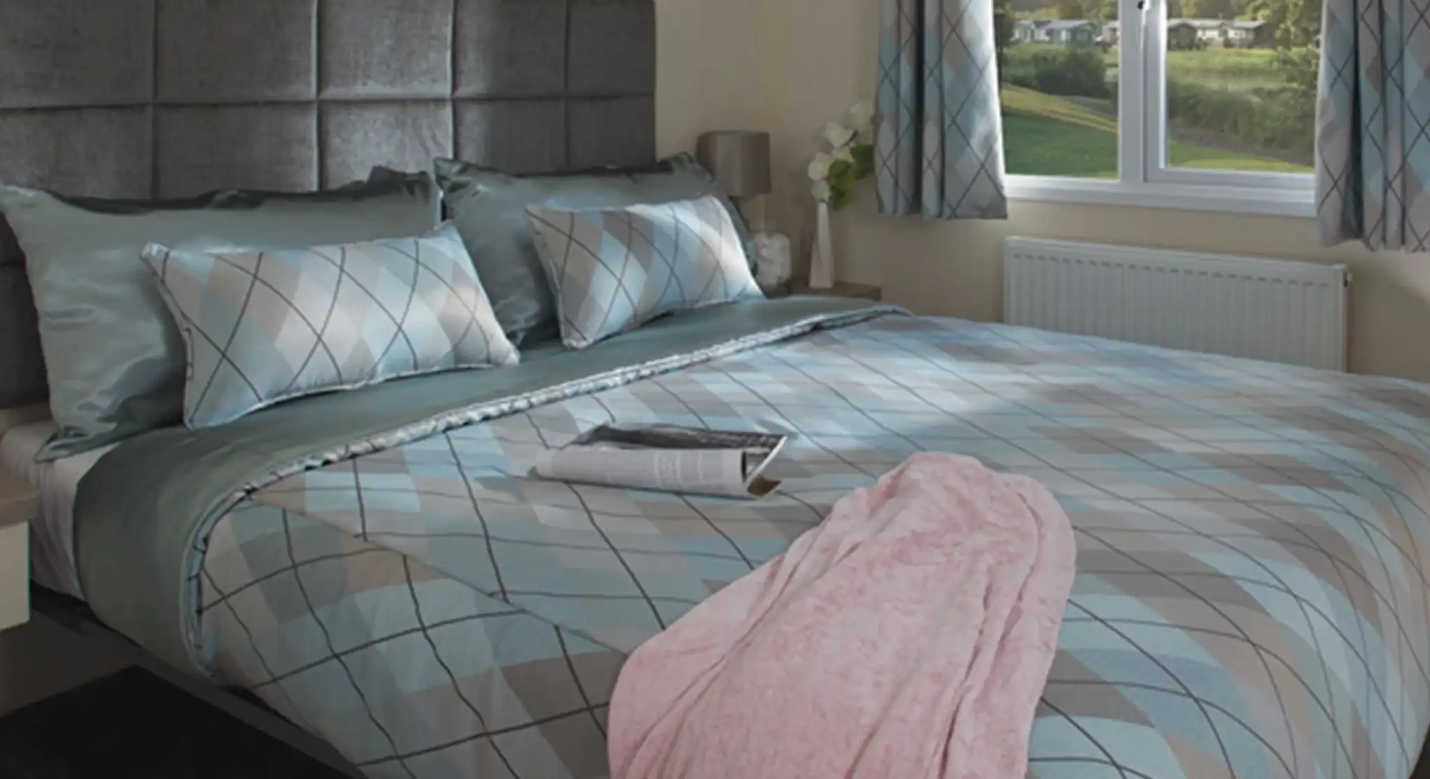 Cosy bedroom with a large bed featuring blue and gray argyle-patterned bedding, a pink blanket, and a magazine. Soft light streams through a window overlooking greenery.