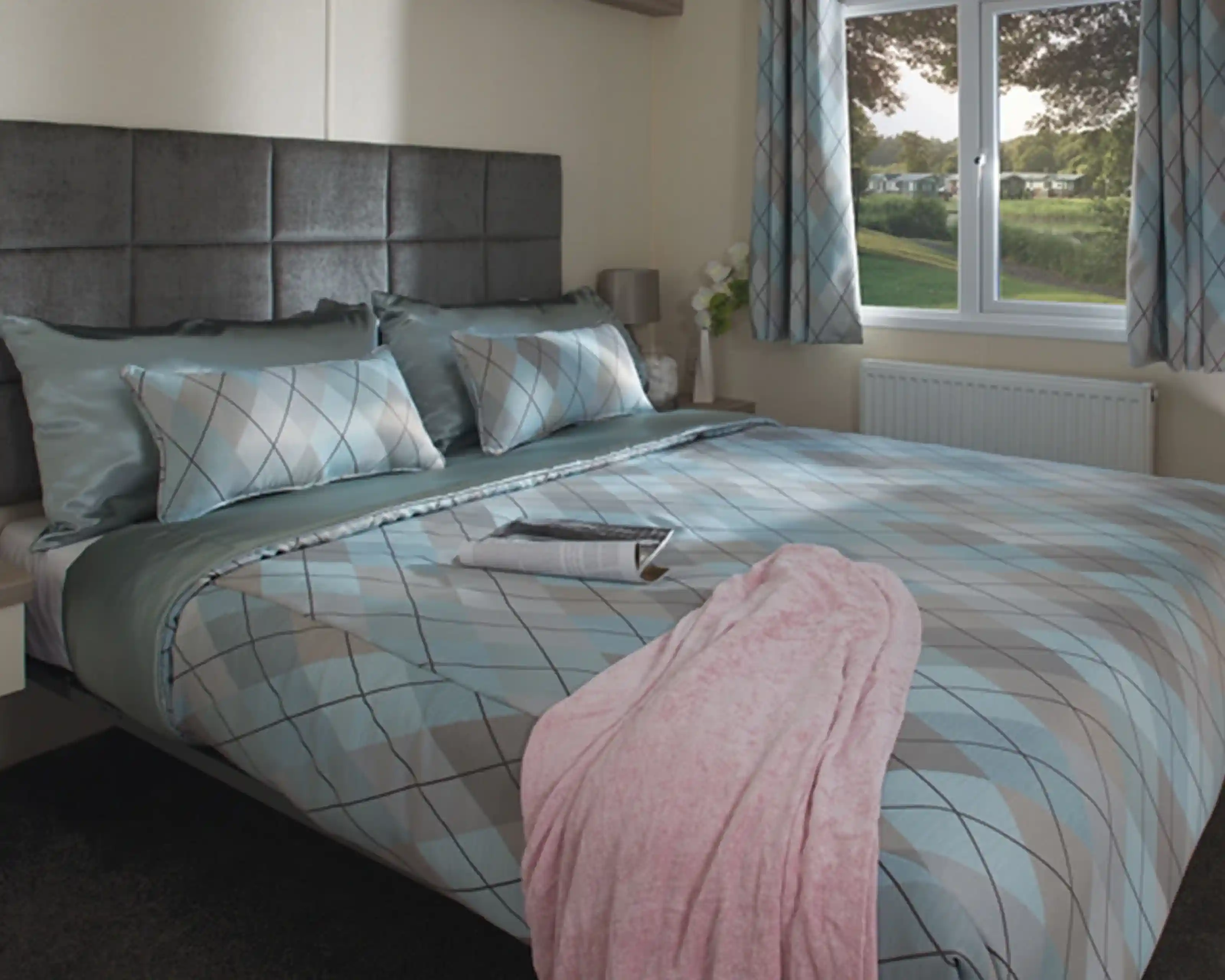 Cosy bedroom with a large bed featuring blue and gray argyle-patterned bedding, a pink blanket, and a magazine. Soft light streams through a window overlooking greenery.