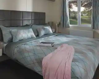Cosy bedroom with a large bed featuring blue and gray argyle-patterned bedding, a pink blanket, and a magazine. Soft light streams through a window overlooking greenery.
