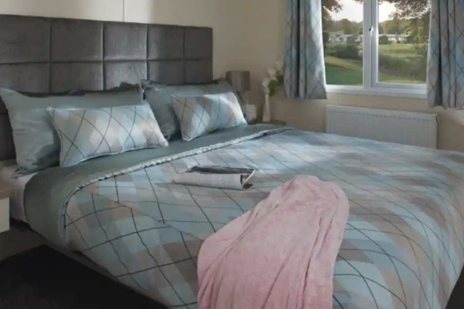 Cosy bedroom with a large bed featuring blue and gray argyle-patterned bedding, a pink blanket, and a magazine. Soft light streams through a window overlooking greenery.