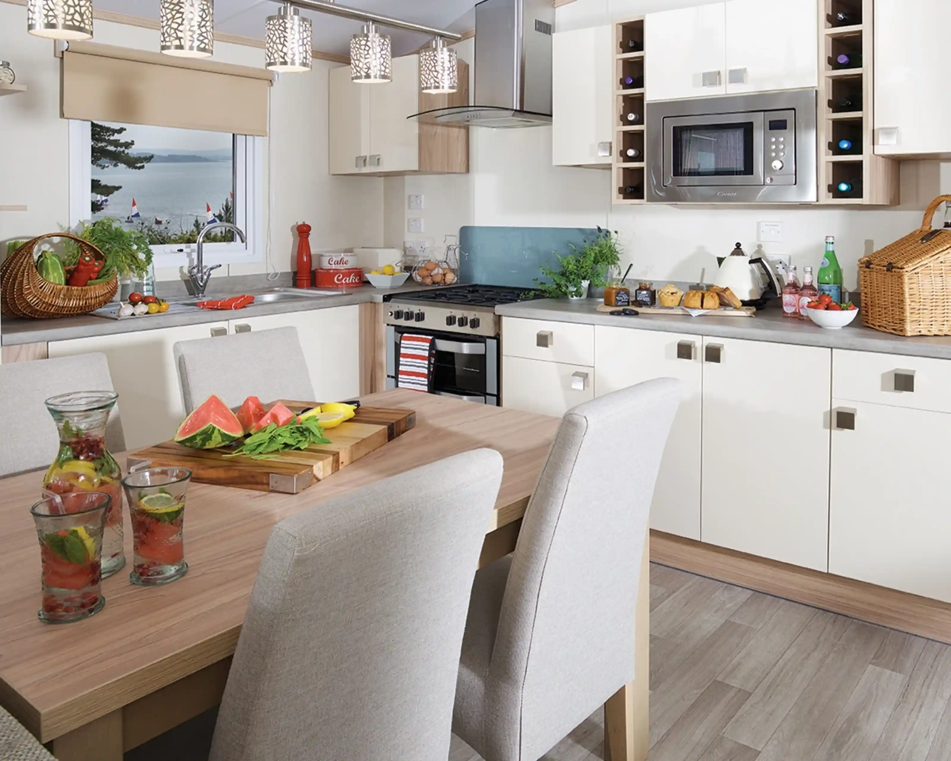 This image captures the bright and modern kitchen and dining area, with an open, airy feel. The kitchen features sleek, contemporary cabinetry in white and light wood tones, with modern appliances like a microwave, oven, and kettle. There's a wine rack built into the upper cabinets and fresh ingredients on the counter, including a basket of vegetables, fruits, and some fresh herbs. The dining table is set with refreshing drinks and a vibrant, fresh watermelon and lemon spread, giving the space a welcoming, summery vibe. Large windows offer a view of the outdoors, enhancing the natural light that floods the room. The overall design is clean, functional, and inviting.