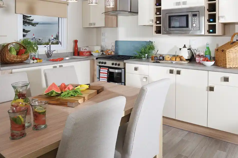 This image captures the bright and modern kitchen and dining area, with an open, airy feel. The kitchen features sleek, contemporary cabinetry in white and light wood tones, with modern appliances like a microwave, oven, and kettle. There's a wine rack built into the upper cabinets and fresh ingredients on the counter, including a basket of vegetables, fruits, and some fresh herbs. The dining table is set with refreshing drinks and a vibrant, fresh watermelon and lemon spread, giving the space a welcoming, summery vibe. Large windows offer a view of the outdoors, enhancing the natural light that floods the room. The overall design is clean, functional, and inviting.