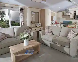 This image showcases a cozy and welcoming lounge area with a light, neutral color scheme. The space features two comfortable sofas adorned with soft, checkered cushions. A wooden coffee table sits between the sofas, decorated with fresh flowers in a vase, adding a touch of warmth and nature to the room. The large windows allow plenty of natural light to fill the space, offering a view of the outdoors, with trees and a glimpse of water visible. Behind the seating area, you can see the open-concept kitchen and dining space, which is bright and modern. The overall vibe is serene, stylish, and perfect for relaxation and socializing.