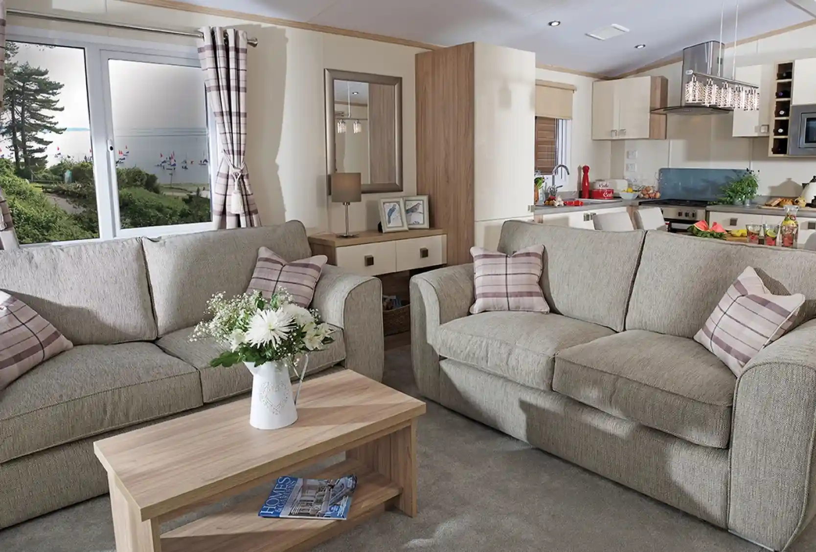 This image showcases a cozy and welcoming lounge area with a light, neutral color scheme. The space features two comfortable sofas adorned with soft, checkered cushions. A wooden coffee table sits between the sofas, decorated with fresh flowers in a vase, adding a touch of warmth and nature to the room. The large windows allow plenty of natural light to fill the space, offering a view of the outdoors, with trees and a glimpse of water visible. Behind the seating area, you can see the open-concept kitchen and dining space, which is bright and modern. The overall vibe is serene, stylish, and perfect for relaxation and socializing.