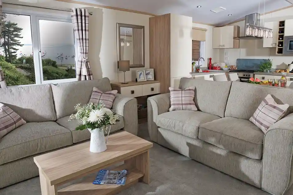 This image showcases a cozy and welcoming lounge area with a light, neutral color scheme. The space features two comfortable sofas adorned with soft, checkered cushions. A wooden coffee table sits between the sofas, decorated with fresh flowers in a vase, adding a touch of warmth and nature to the room. The large windows allow plenty of natural light to fill the space, offering a view of the outdoors, with trees and a glimpse of water visible. Behind the seating area, you can see the open-concept kitchen and dining space, which is bright and modern. The overall vibe is serene, stylish, and perfect for relaxation and socializing.