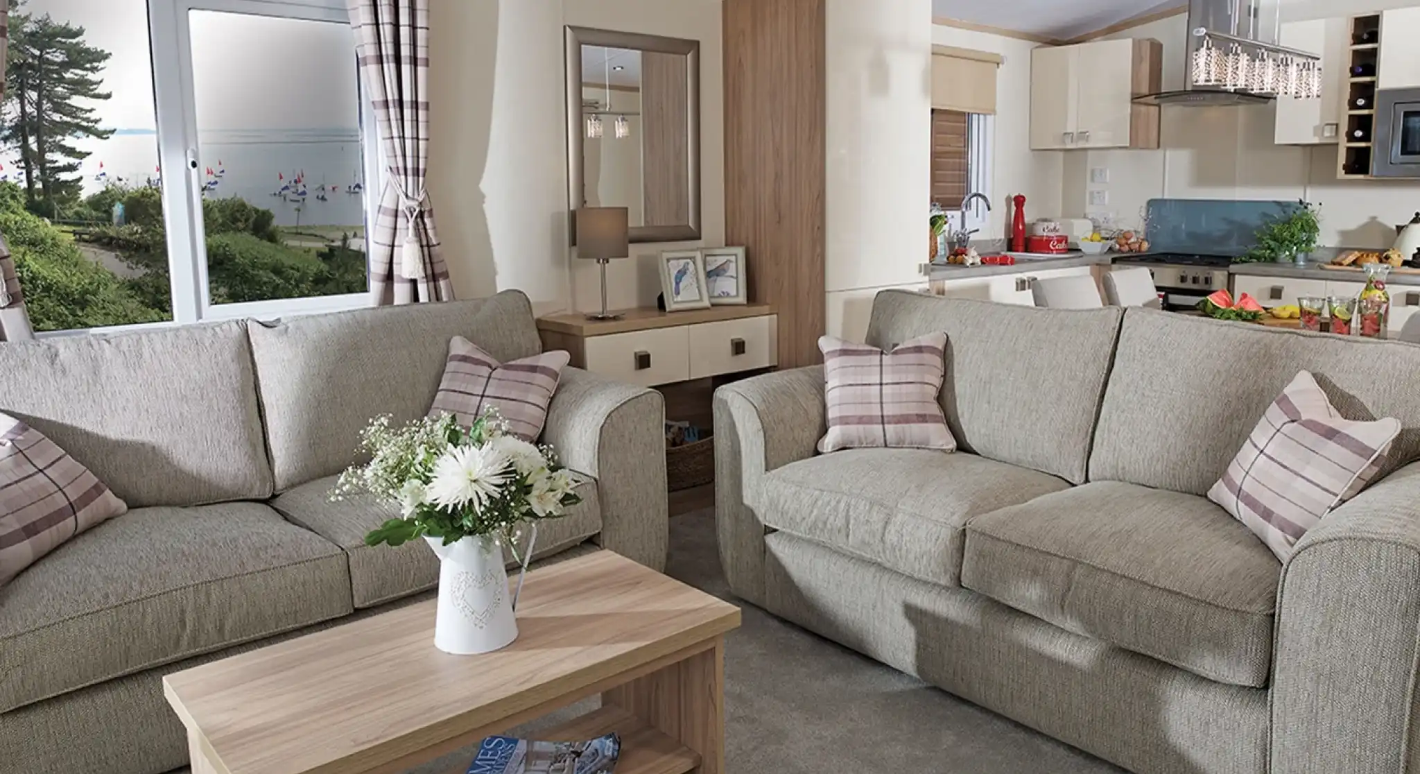 This image showcases a cozy and welcoming lounge area with a light, neutral color scheme. The space features two comfortable sofas adorned with soft, checkered cushions. A wooden coffee table sits between the sofas, decorated with fresh flowers in a vase, adding a touch of warmth and nature to the room. The large windows allow plenty of natural light to fill the space, offering a view of the outdoors, with trees and a glimpse of water visible. Behind the seating area, you can see the open-concept kitchen and dining space, which is bright and modern. The overall vibe is serene, stylish, and perfect for relaxation and socializing.