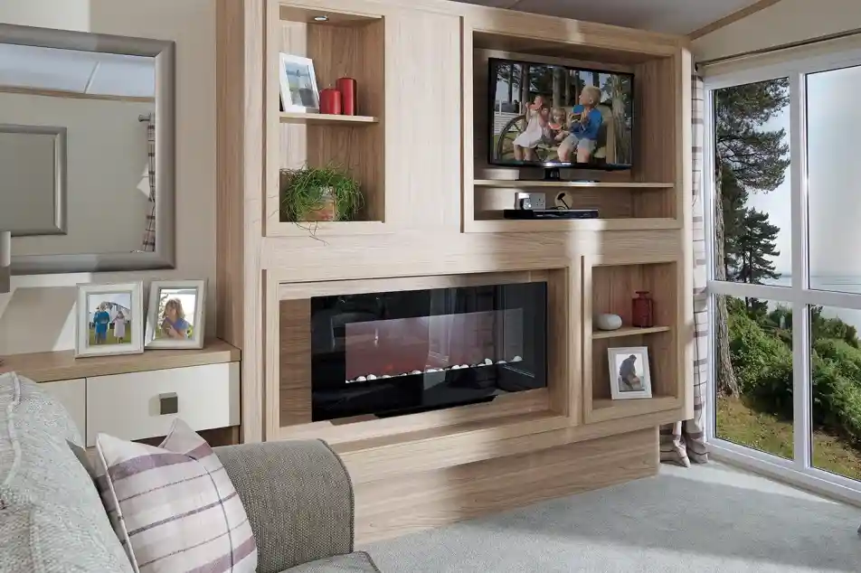 This image showcases the other side of the same living room, emphasizing the sleek, modern entertainment and storage unit. The wood paneling matches the neutral tones of the room, and the built-in television is perched on a shelf, with family photos and decorative items surrounding it. Below the TV, there's a contemporary electric fireplace that adds a cozy ambiance. The large mirror on the left reflects light, making the room feel even more spacious. The overall design continues with the light, airy color scheme and tranquil feel, while maintaining a balance between functionality and comfort.