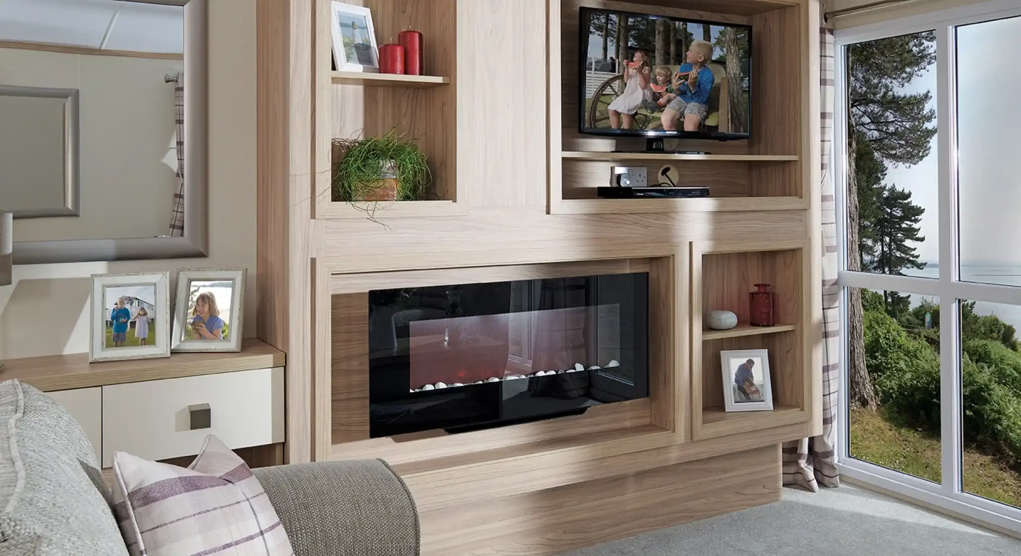 This image showcases the other side of the same living room, emphasizing the sleek, modern entertainment and storage unit. The wood paneling matches the neutral tones of the room, and the built-in television is perched on a shelf, with family photos and decorative items surrounding it. Below the TV, there's a contemporary electric fireplace that adds a cozy ambiance. The large mirror on the left reflects light, making the room feel even more spacious. The overall design continues with the light, airy color scheme and tranquil feel, while maintaining a balance between functionality and comfort.