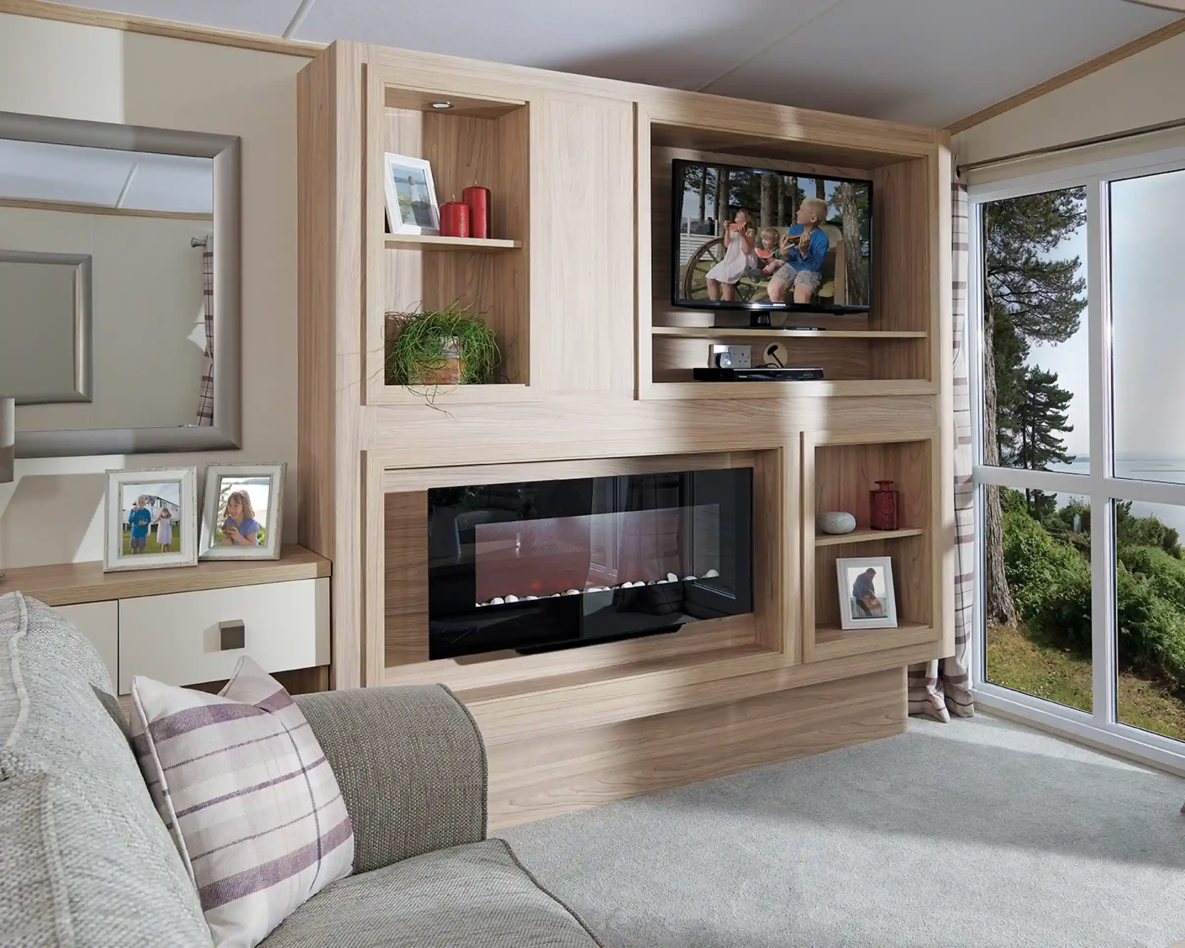 This image showcases the other side of the same living room, emphasizing the sleek, modern entertainment and storage unit. The wood paneling matches the neutral tones of the room, and the built-in television is perched on a shelf, with family photos and decorative items surrounding it. Below the TV, there's a contemporary electric fireplace that adds a cozy ambiance. The large mirror on the left reflects light, making the room feel even more spacious. The overall design continues with the light, airy color scheme and tranquil feel, while maintaining a balance between functionality and comfort.