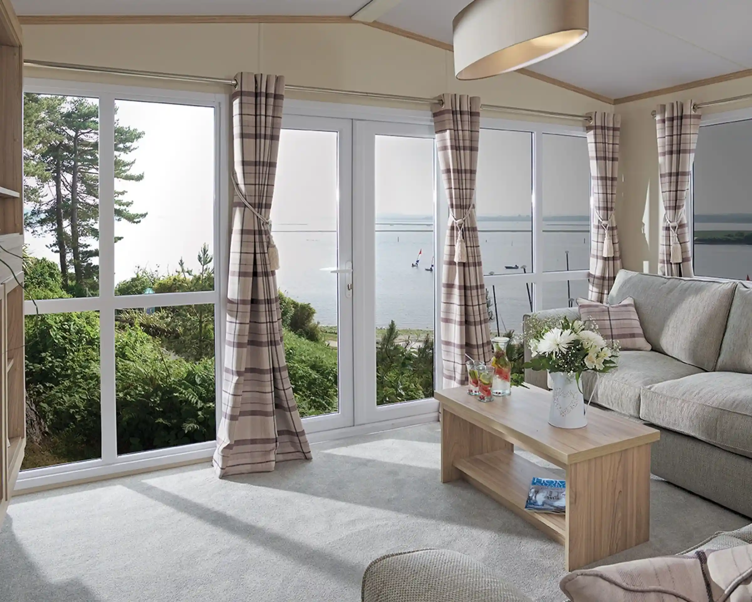 The image shows a cosy, modern living room with a soft, neutral color scheme. The room has large windows, providing a beautiful view of the outdoors, possibly a lake or river, with sailboats visible in the distance. The furniture includes a comfortable sofa with cushions and a simple wooden coffee table, adorned with a flower arrangement and drinks. The curtains are checked in a subtle, pastel color, and the light from the large window creates a bright and airy atmosphere. The space feels inviting, tranquil, and connected to nature outside.