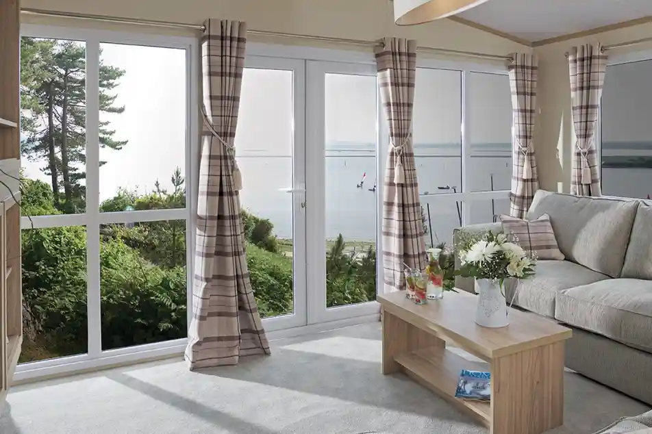 The image shows a cosy, modern living room with a soft, neutral color scheme. The room has large windows, providing a beautiful view of the outdoors, possibly a lake or river, with sailboats visible in the distance. The furniture includes a comfortable sofa with cushions and a simple wooden coffee table, adorned with a flower arrangement and drinks. The curtains are checked in a subtle, pastel color, and the light from the large window creates a bright and airy atmosphere. The space feels inviting, tranquil, and connected to nature outside.