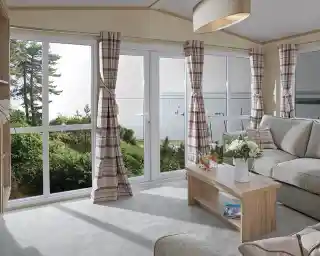 The image shows a cosy, modern living room with a soft, neutral color scheme. The room has large windows, providing a beautiful view of the outdoors, possibly a lake or river, with sailboats visible in the distance. The furniture includes a comfortable sofa with cushions and a simple wooden coffee table, adorned with a flower arrangement and drinks. The curtains are checked in a subtle, pastel color, and the light from the large window creates a bright and airy atmosphere. The space feels inviting, tranquil, and connected to nature outside.